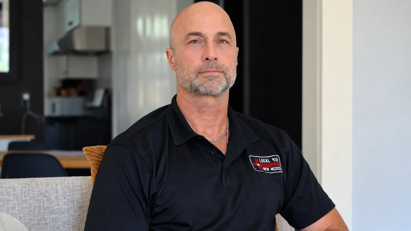 A bald man with a gray beard, wearing a black polo shirt, sits on a gray couch in a bright living room and looks directly at the camera.
