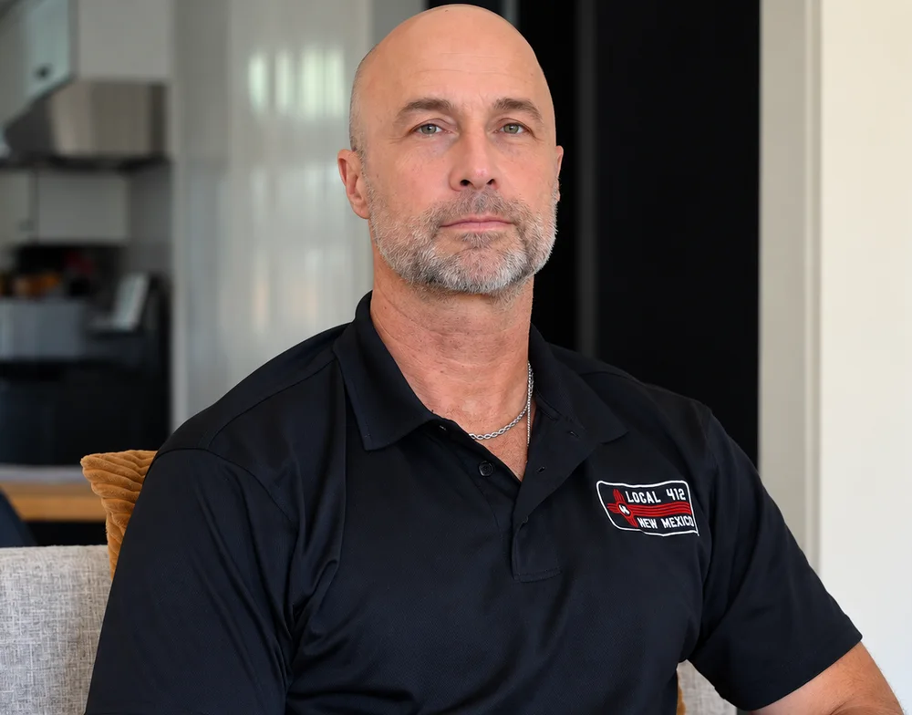 A bald man with a gray beard, wearing a black polo shirt, sits on a gray couch and looks directly at the camera.